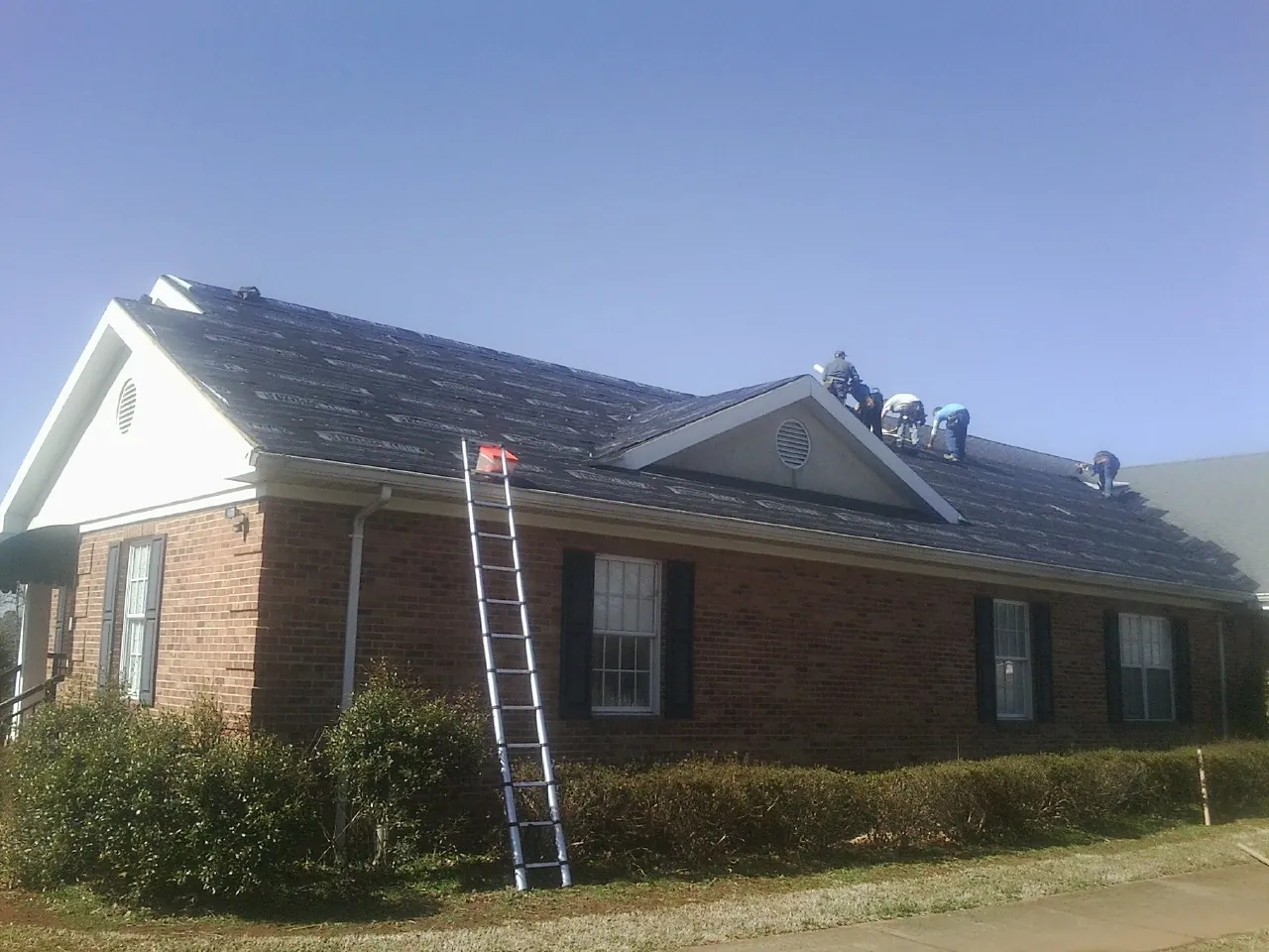 Crew tearing shingles off a brick ranch roof with a ladder set — Emergency Roof Repair work by our crew in Hahira